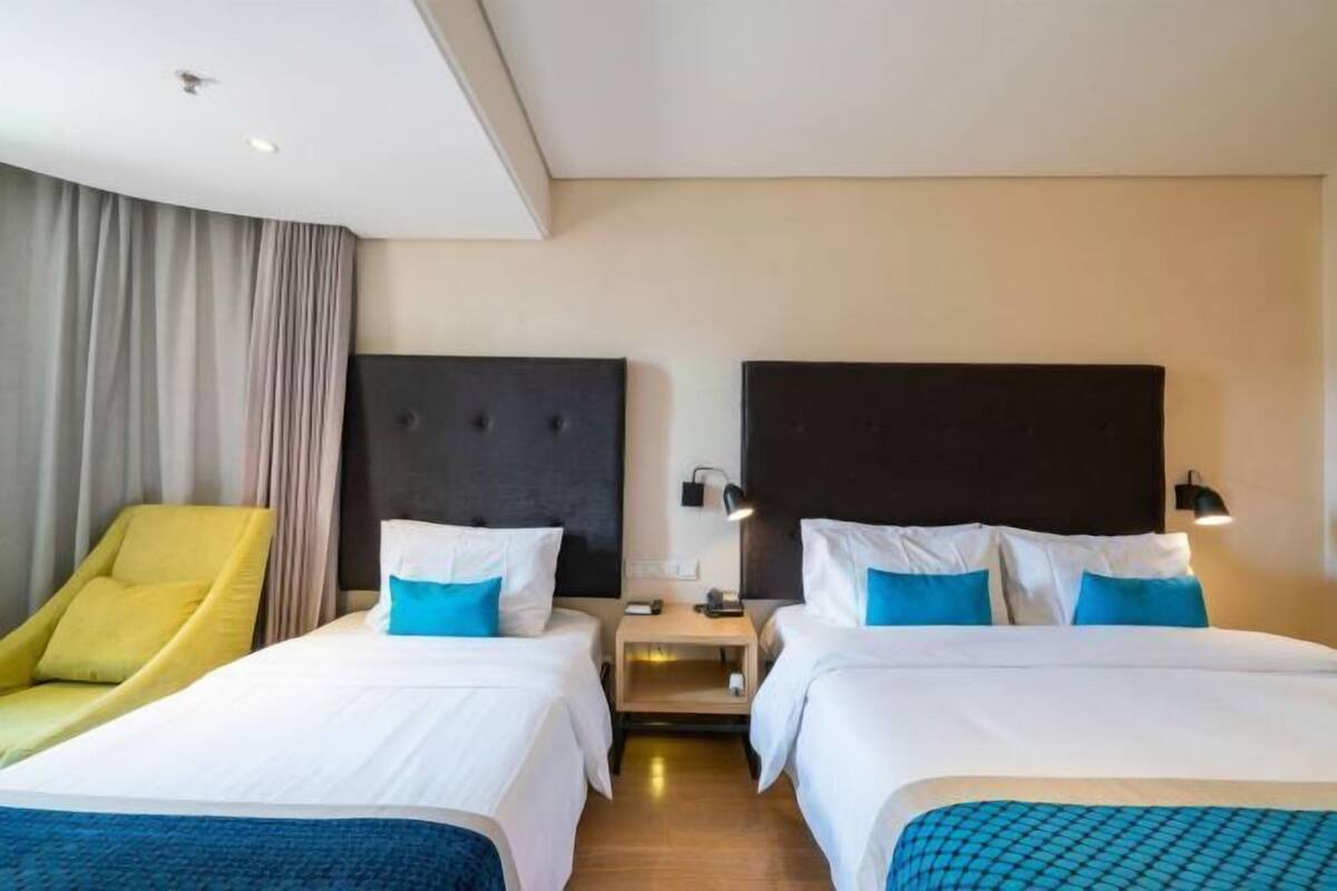 A hotel room with two beds, a yellow armchair, and a nightstand with a lamp.