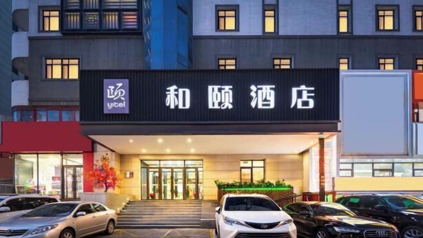 A hotel entrance with a sign in Chinese characters, a parking lot with several cars, and a building facade with multiple windows.