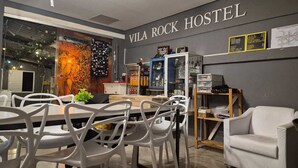 En modern vandrarhemsinredning med en matplats, vita stolar, en soffa och en vägg med namnet ”VILA ROCK HOSTEL”.