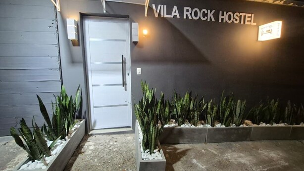 En hostelentrén med en skylt som visar texten ”VILA ROCK HOSTEL”, en dörr och planteringskärl med gröna växter.