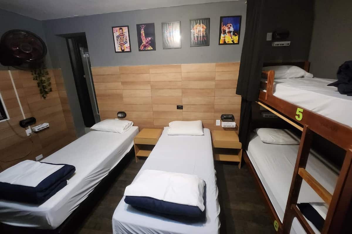 Ein Zimmer mit drei Einzelbetten, einem Holzkopfteil, einem Wandventilator und mehreren gerahmten Bildern.