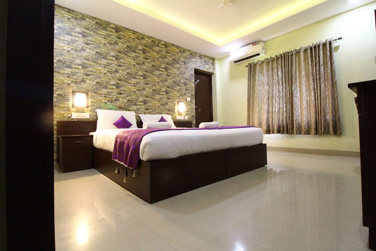 Un dormitorio moderno con pared de piedra, una cama grande con ropa de cama morada y una mesita de noche con lámpara.