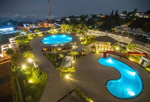 Un resort con una amplia piscina, zonas de descanso y vistas de la ciudad iluminada de noche.
