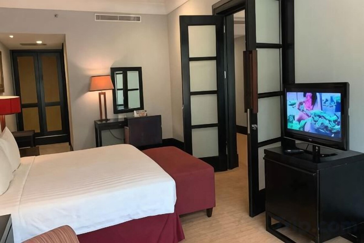 A hotel room with a bed, a television, a desk with a lamp, and a wardrobe.