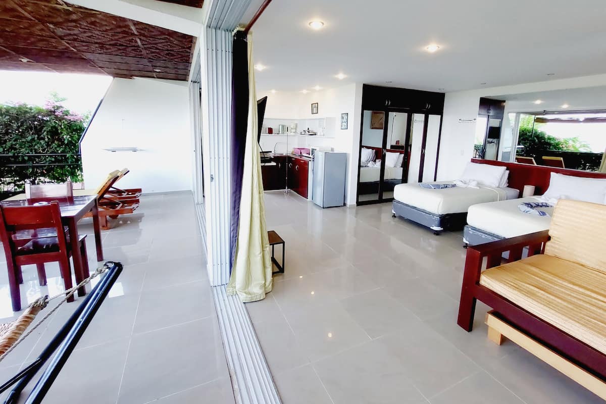 A modern hotel room with a balcony, a dining area with wooden furniture, a kitchen area, and a bed with a suitcase.