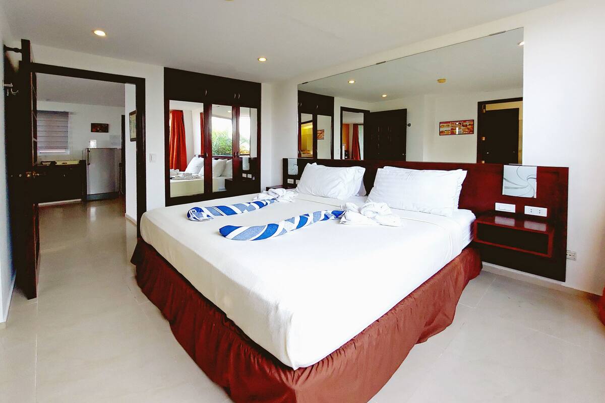 A large bed with white linens, a red skirted base, and blue striped towels. There is a wooden nightstand with a mirror and a small table. The room has white walls and a large mirror on one wall.