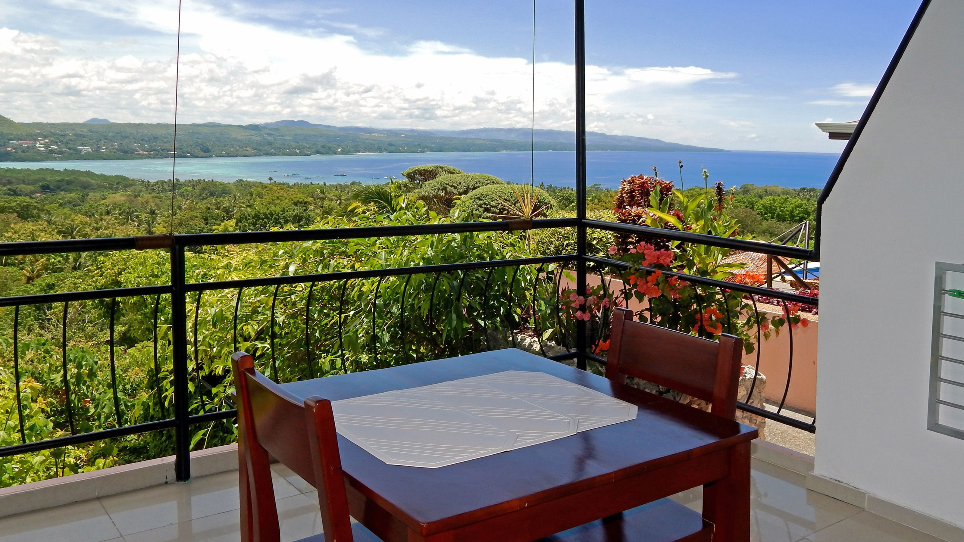 A balcony with a wooden chair and table, overlooking a scenic view of a coastline and lush greenery.
