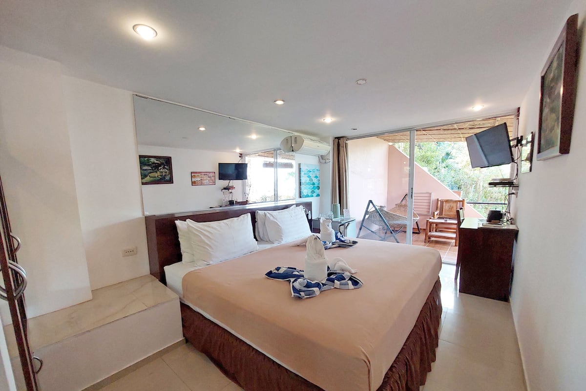 A hotel room with a large bed, a desk with a computer, a TV, and a balcony with a hammock.