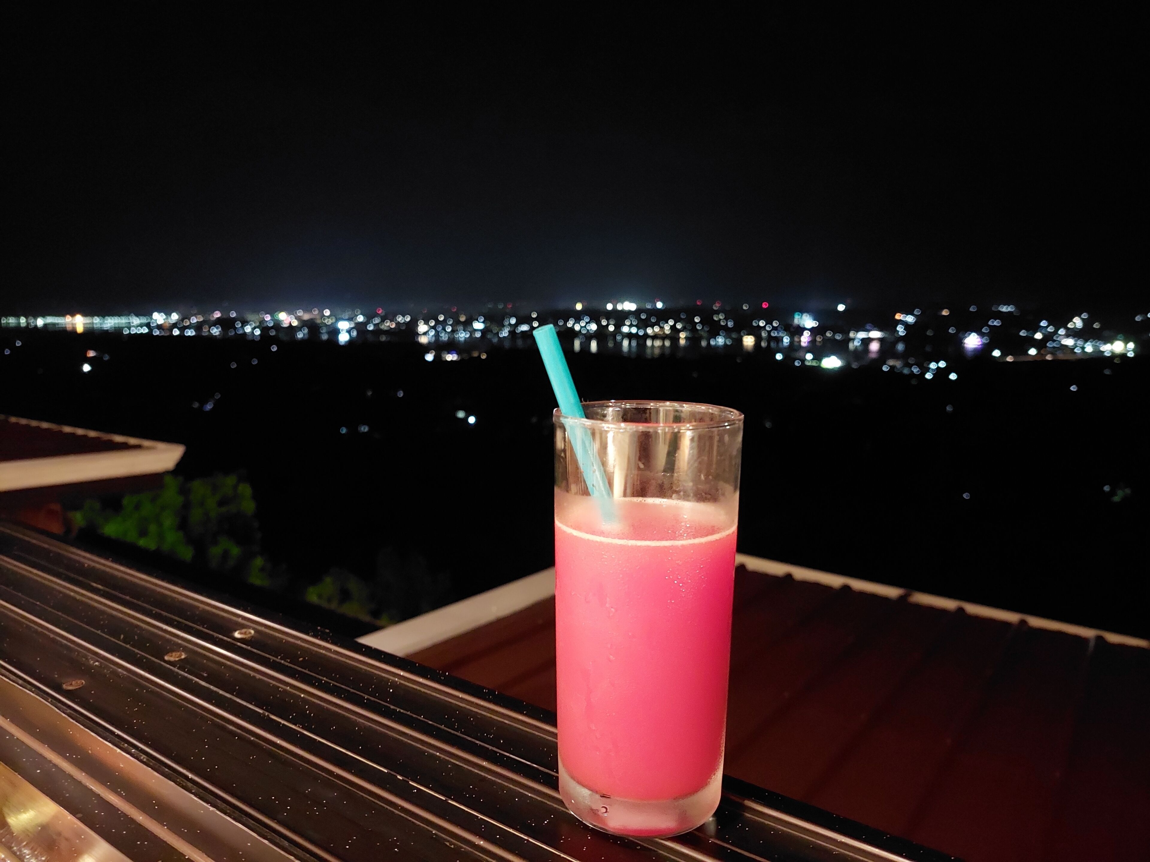 A glass of pink drink with a straw on a balcony with a city view at night.