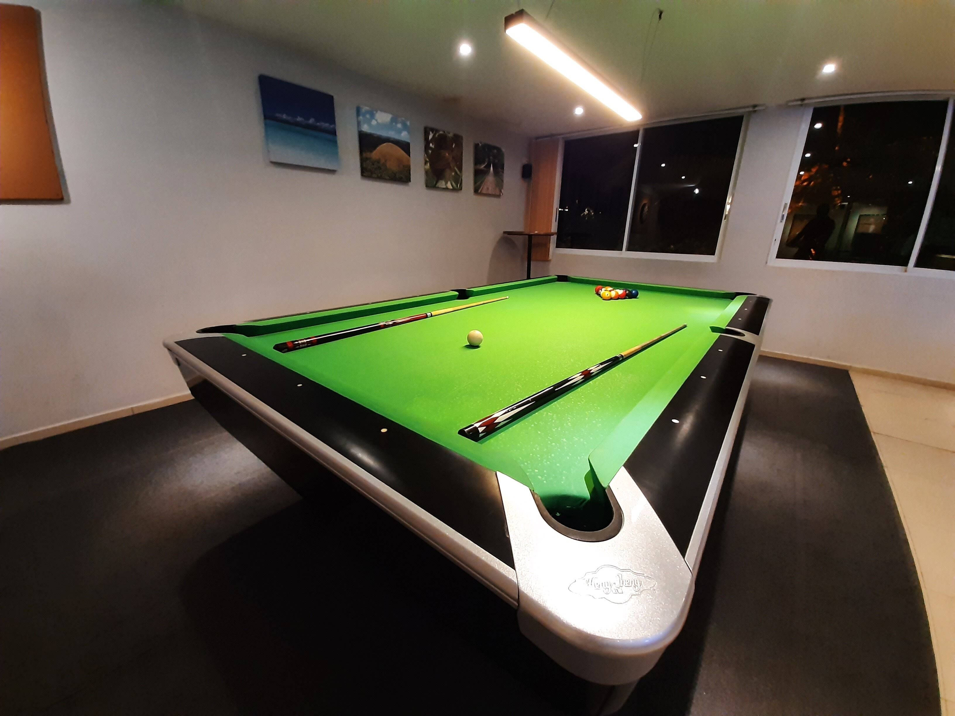 Een pooltafel met een groene viltkleed, omringd door een donkere vloer, in een kamer met witte muren en een raam.