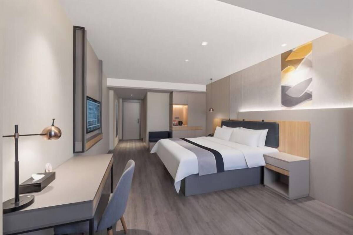 A modern hotel room with a bed, bedside table, desk, chair, and a large window.