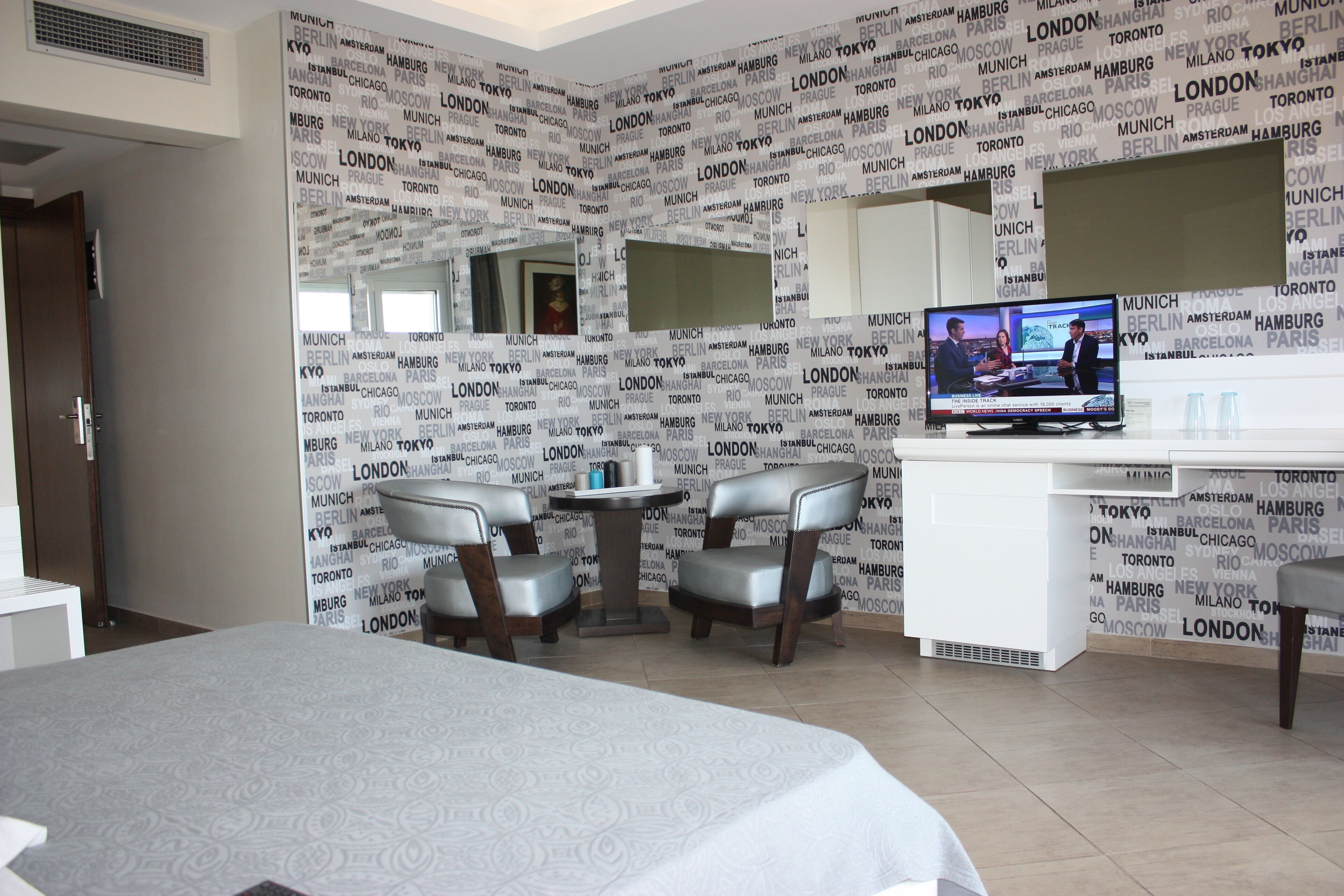 A hotel room with a bed, a desk, two chairs, a television, and a wall with city names.