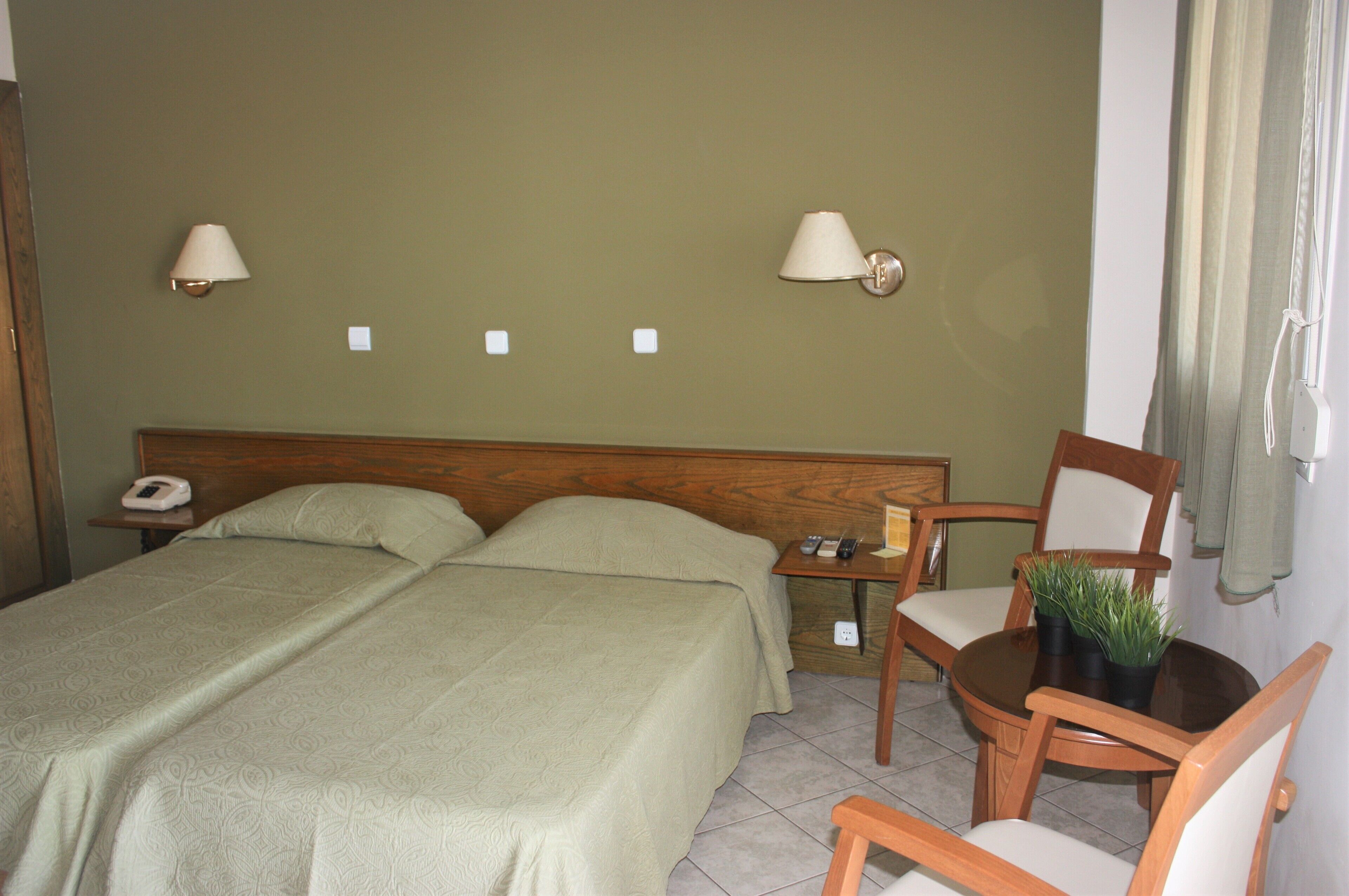Een hotelkamer met twee bedden, een houten hoofdbord, nachtkastjes met lampen, een kleine tafel met stoelen en een potplant.