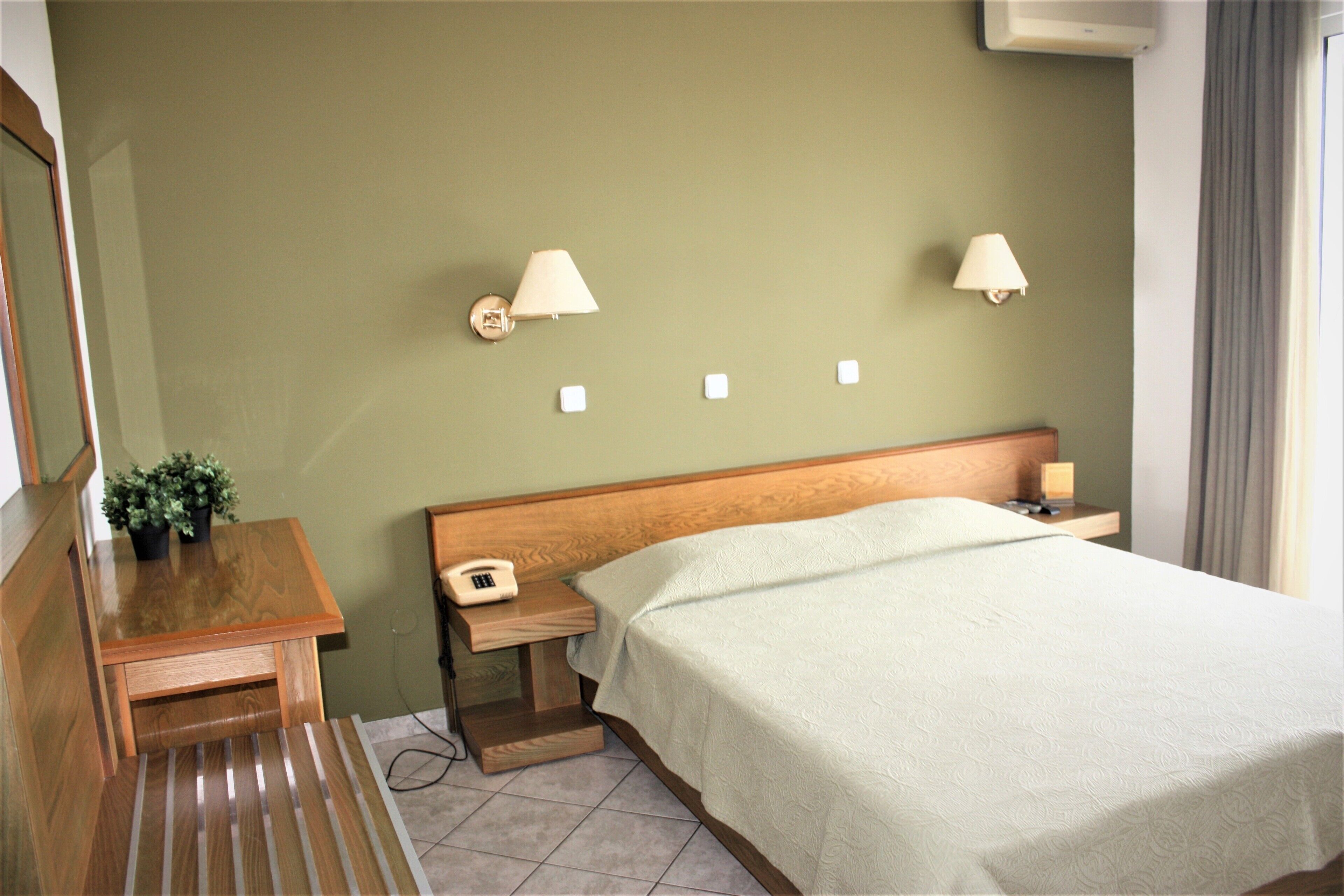 A hotel room with a bed, bedside lamps, a wooden nightstand, a telephone, and a small table with a plant.