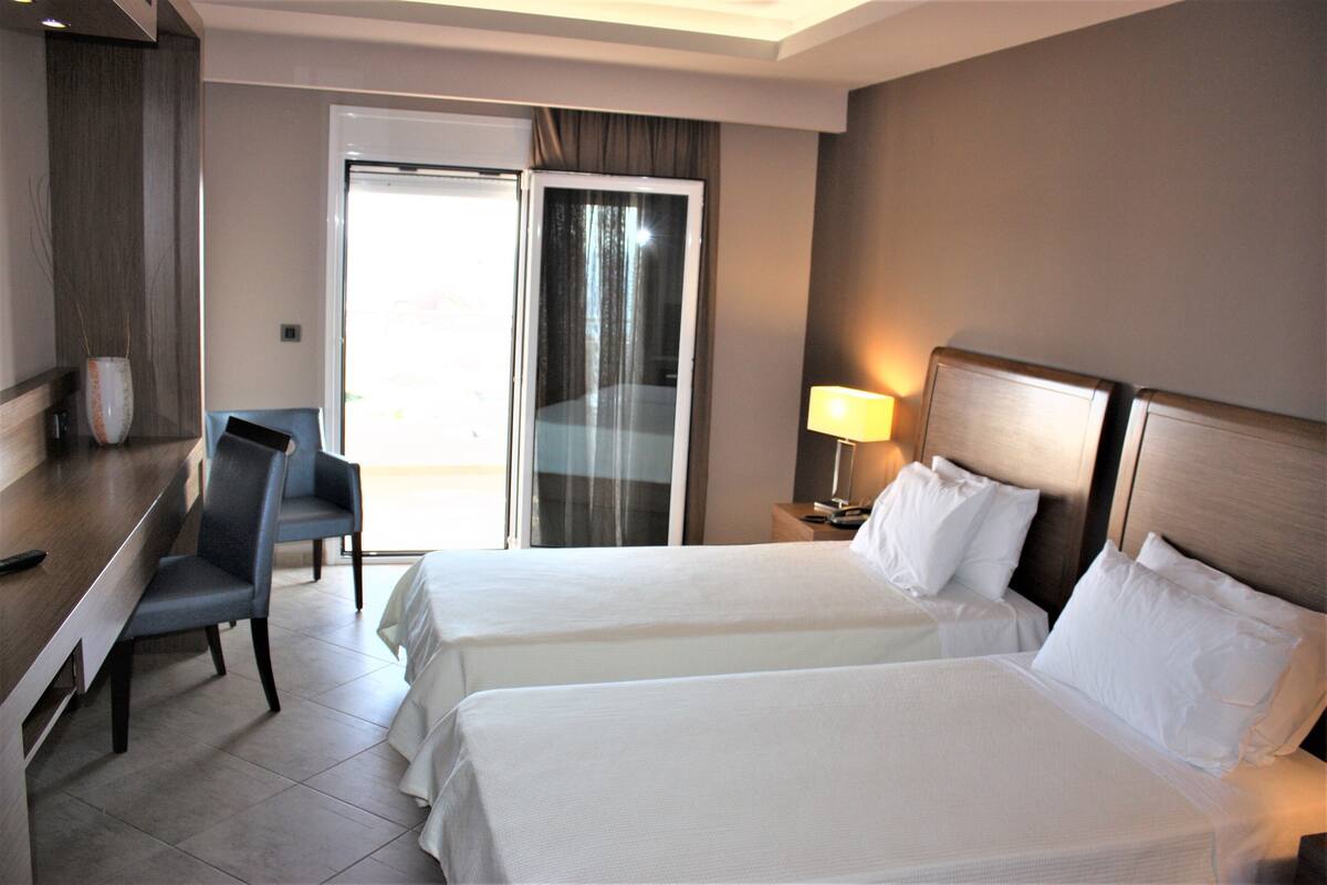 A hotel room with two beds, a desk, a chair, and a sliding glass door to the outside.
