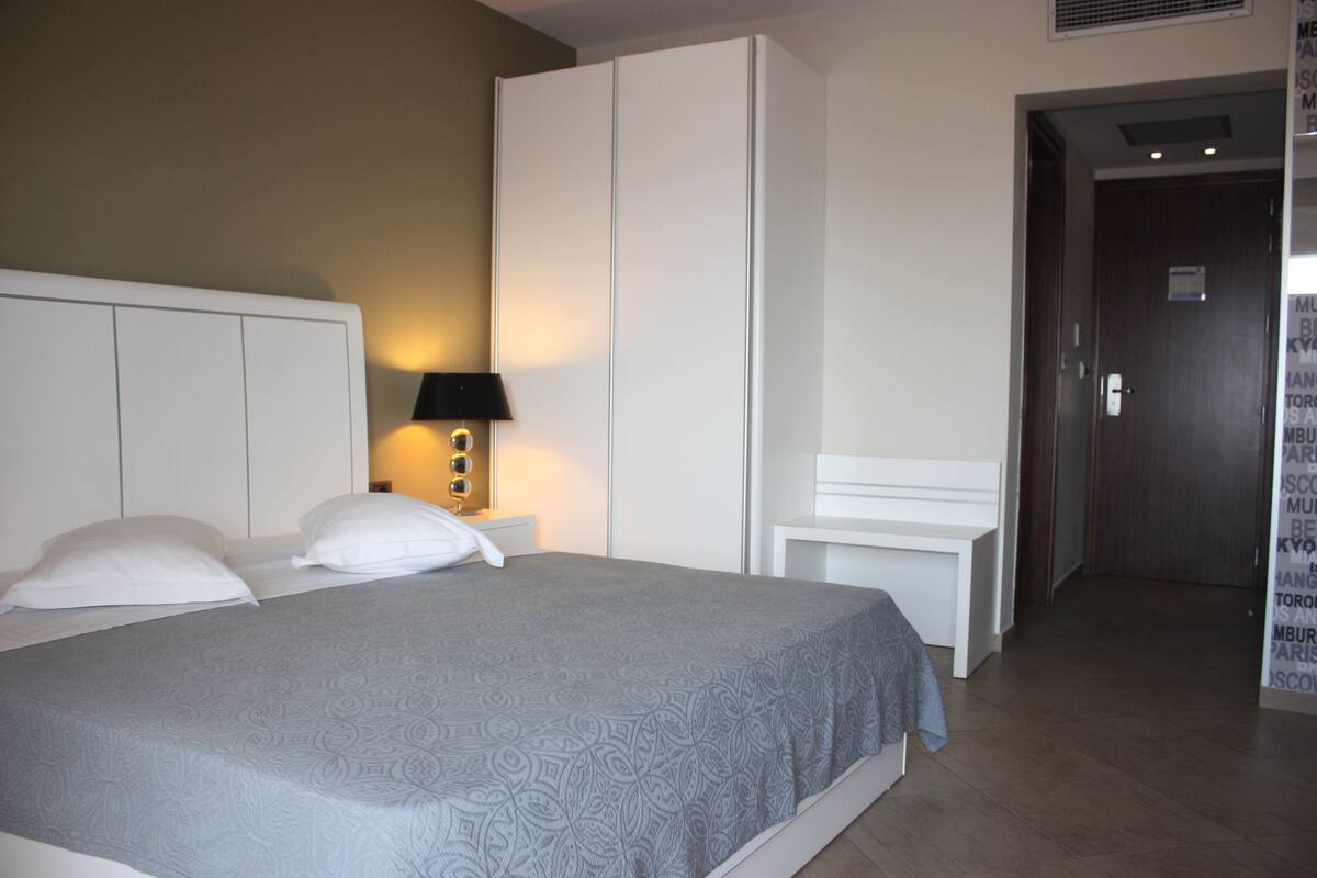 A hotel room with a bed, a bedside table with a lamp, a wardrobe, and a door leading to another room.