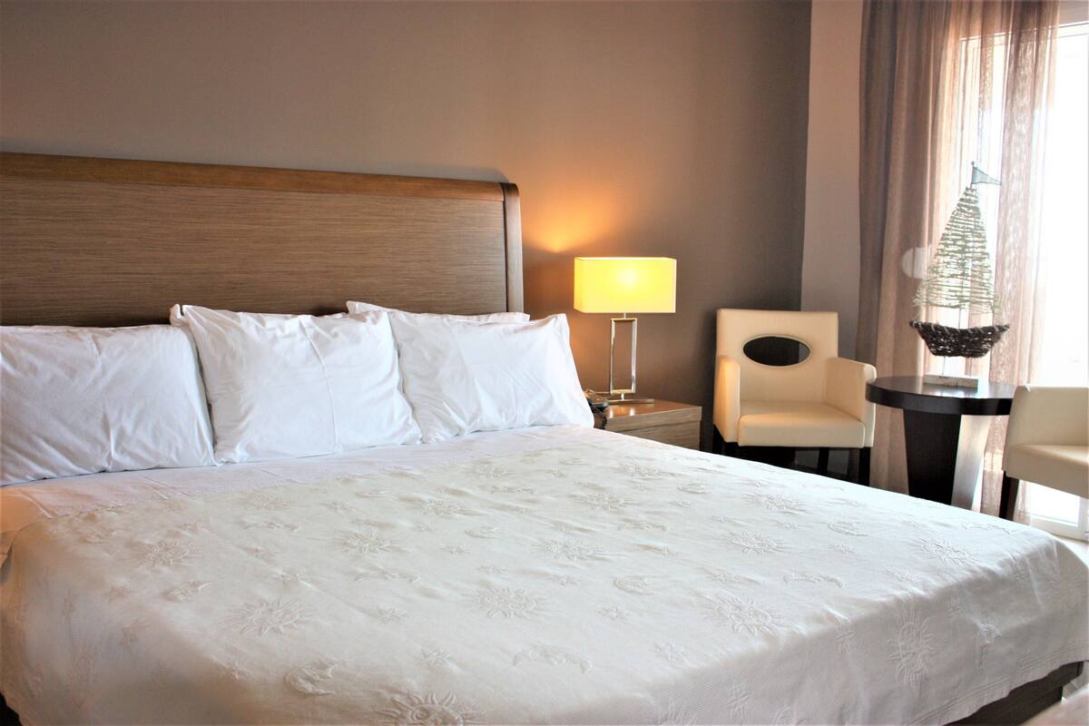 A neatly made bed with white linens, a bedside table with a lamp, a chair, and a decorative item on a small table.