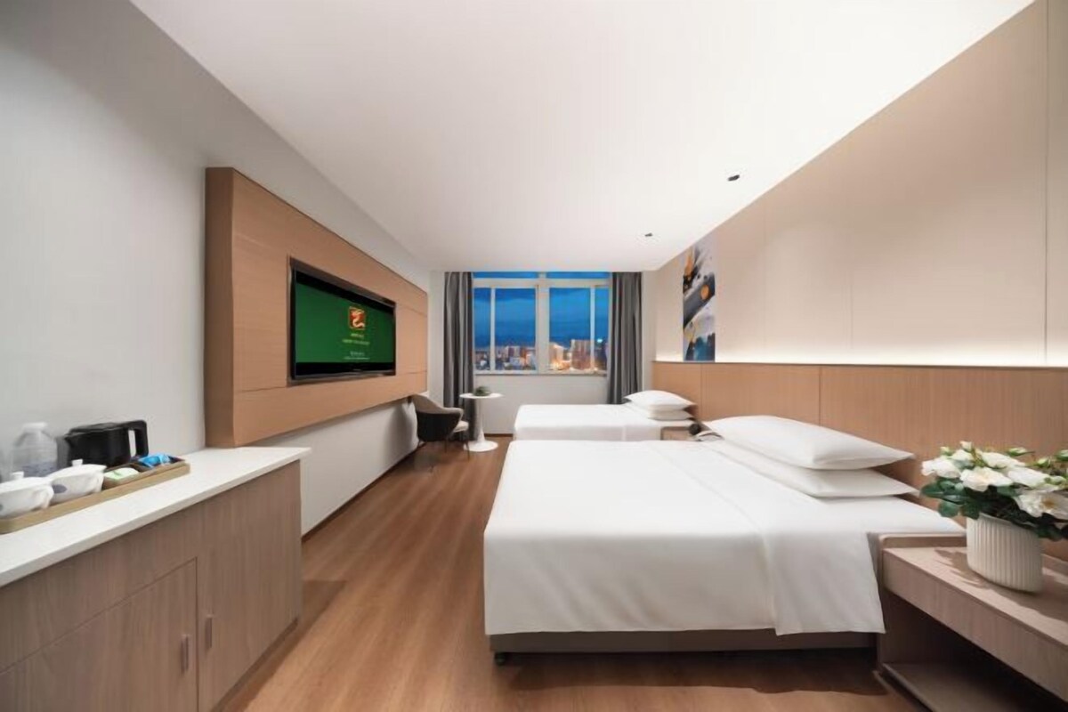 Una habitación de hotel moderna con una cama grande, un televisor montado en la pared, una mesita con una silla y una ventana con vistas a la ciudad.