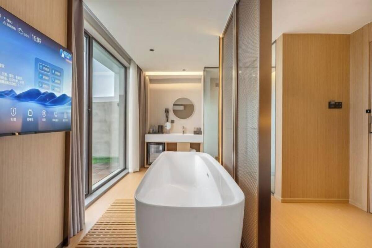 A modern bathroom with a large bathtub, a television mounted on the wall, and a view of the outdoors through the window.