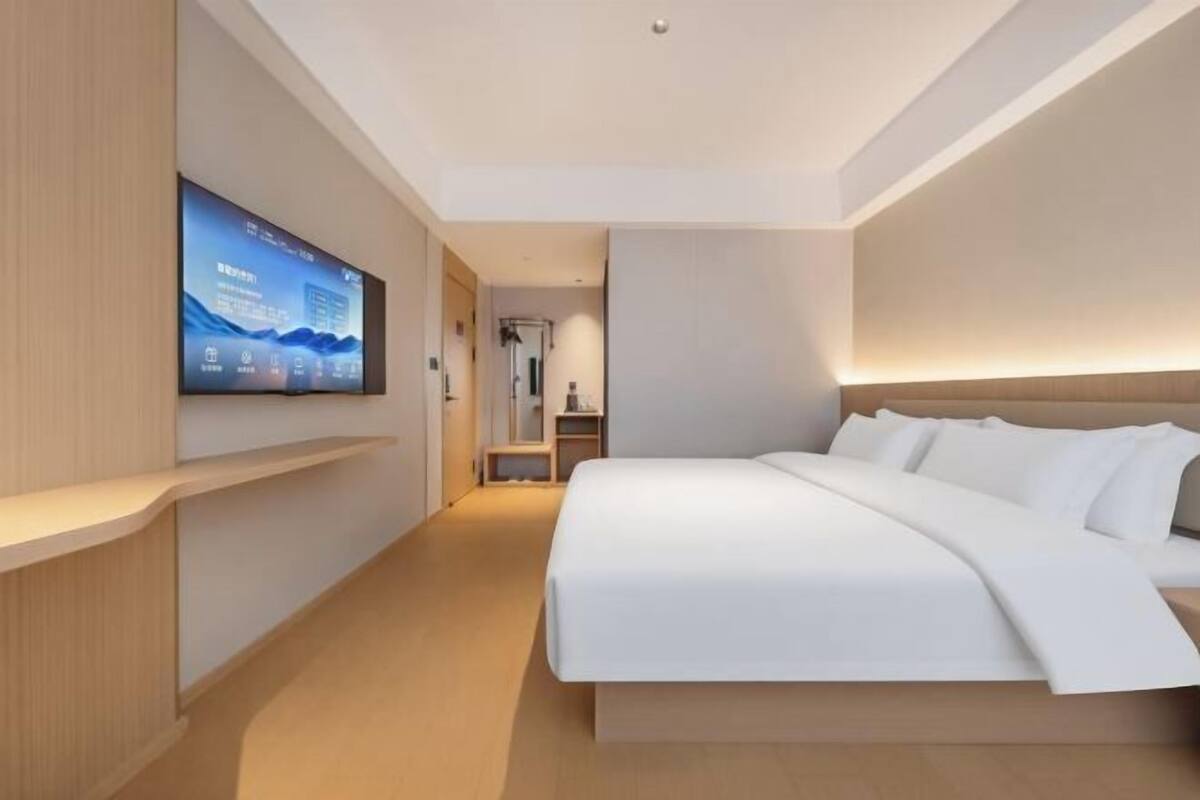A modern hotel room with a large bed, a flat-screen TV mounted on the wall, and a wooden bedside table.