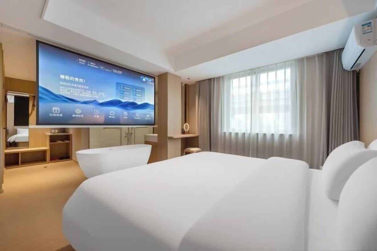A hotel room with a large flat-screen TV, a bed, a desk, and a window with curtains.