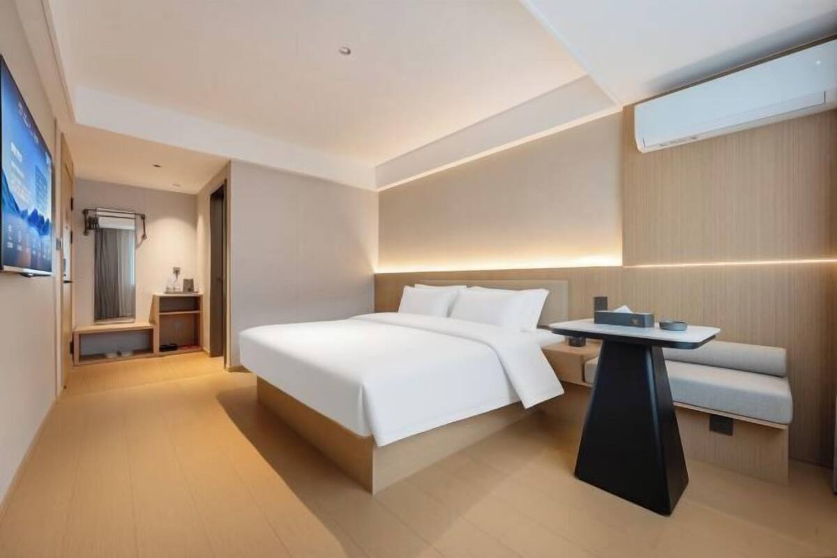 A modern hotel room with a large bed, a bedside table, a built-in bench, and a wall-mounted TV.