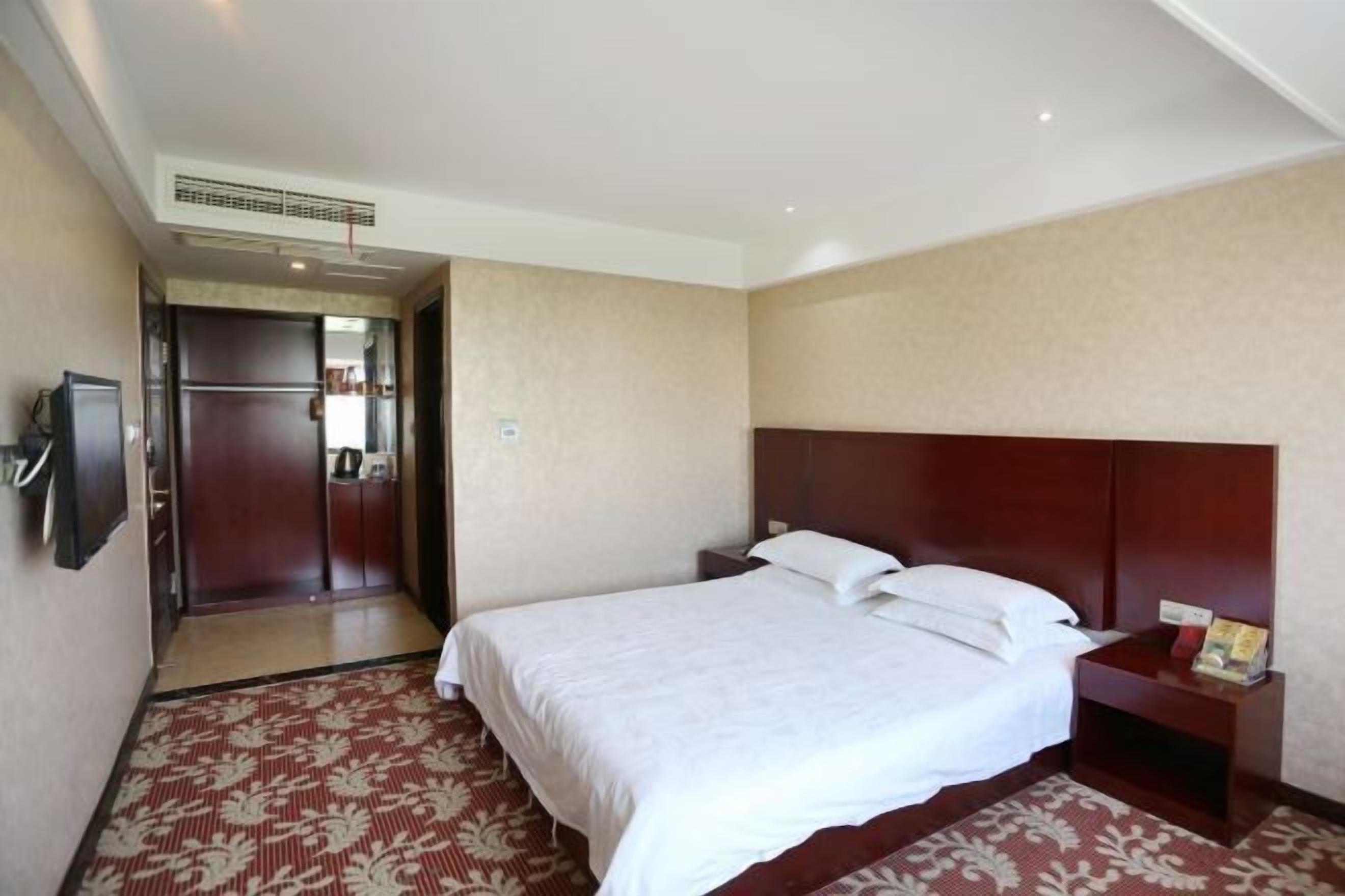 A hotel room with a large bed, a wooden headboard, a bedside table with a lamp, and a carpeted floor.