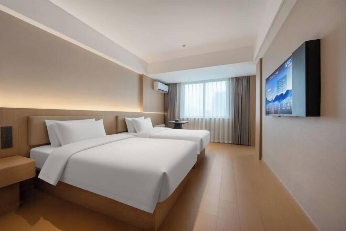 A hotel room with two beds, a TV mounted on the wall, and a window with curtains.