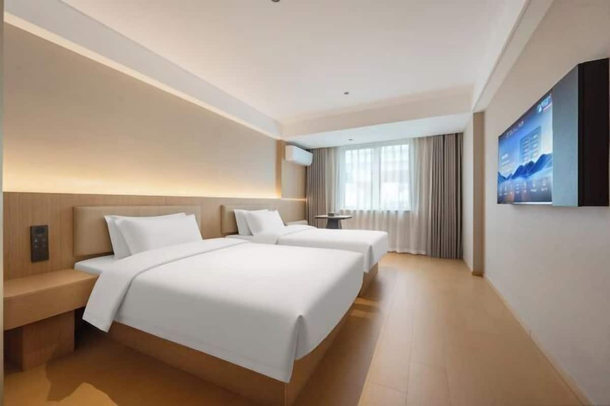 A hotel room with two beds, a bedside table, a desk, a chair, a window with curtains, and a wall-mounted TV.
