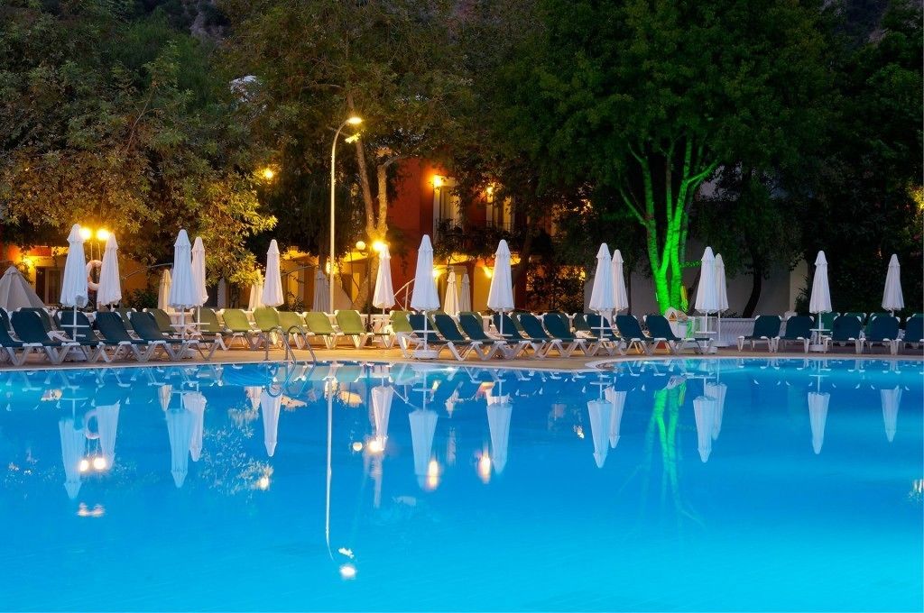 A poolside area with lounge chairs and umbrellas, surrounded by trees and illuminated by ambient lighting.