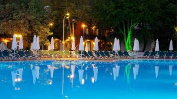 A poolside area with lounge chairs and umbrellas, surrounded by trees and illuminated by ambient lighting.