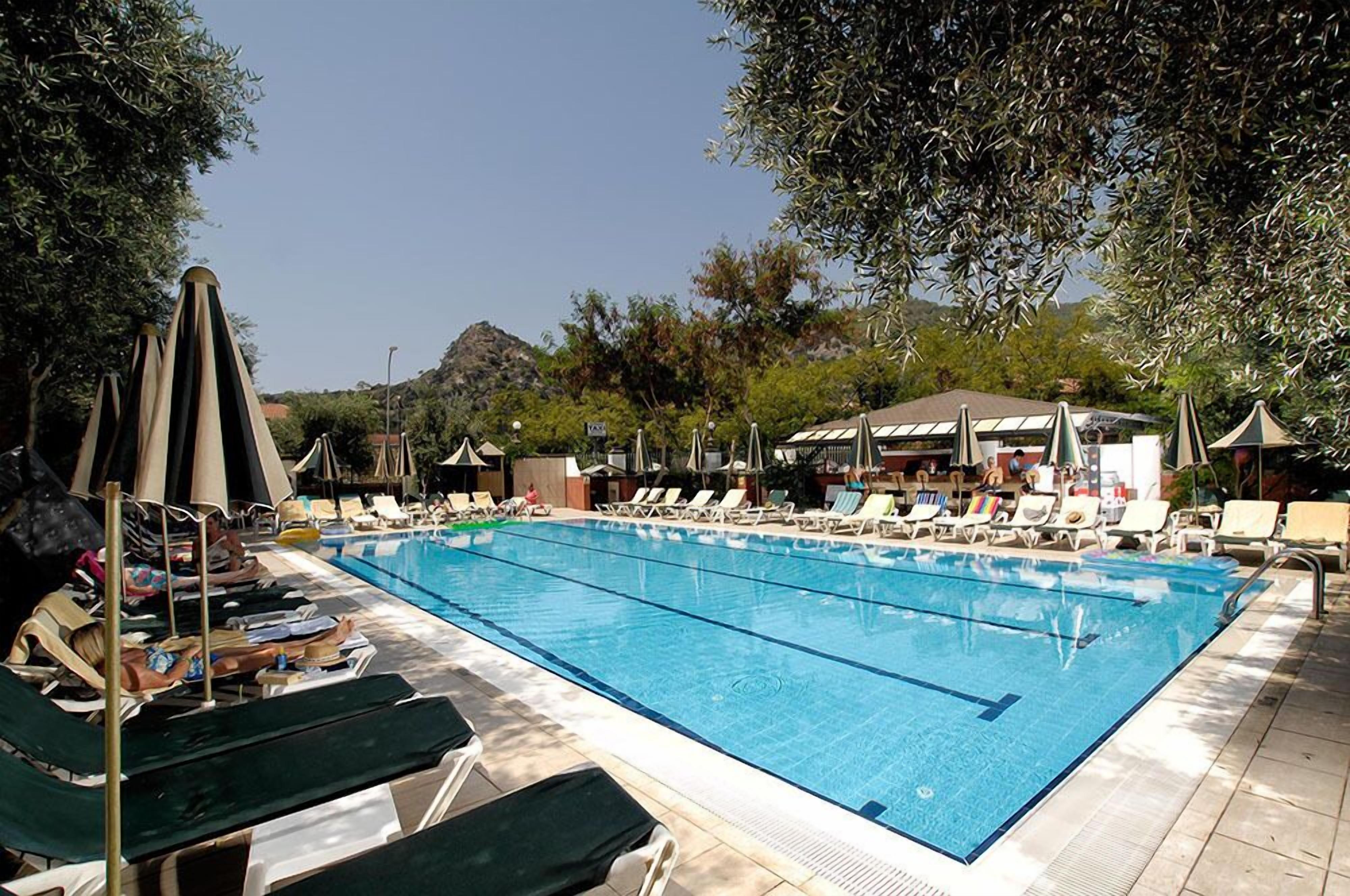 Una piscina con sillas de descanso y sombrillas, rodeada de árboles y un edificio de techo inclinado.