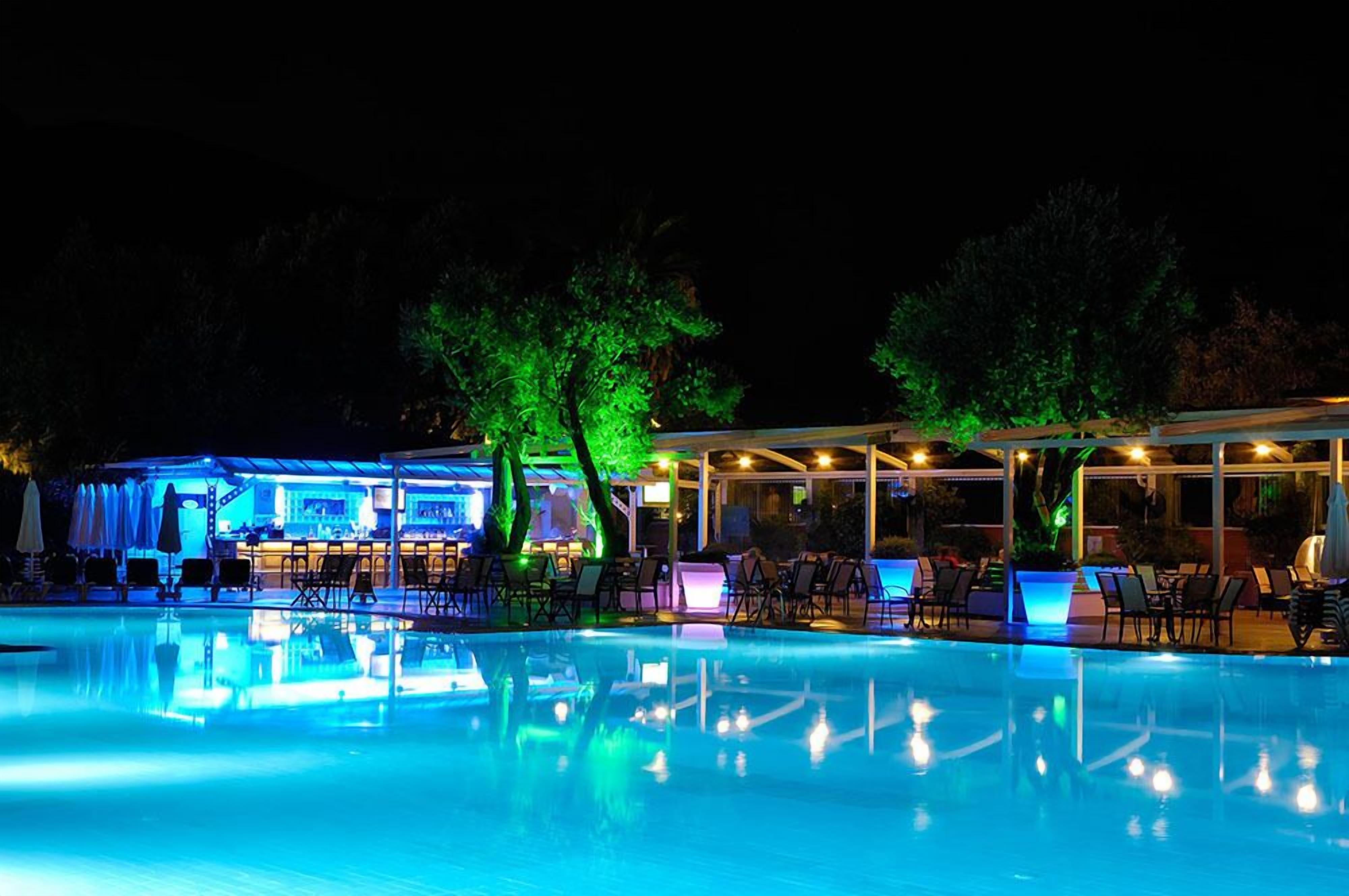 Zona de piscina nocturna con sillones de descanso y un bar.