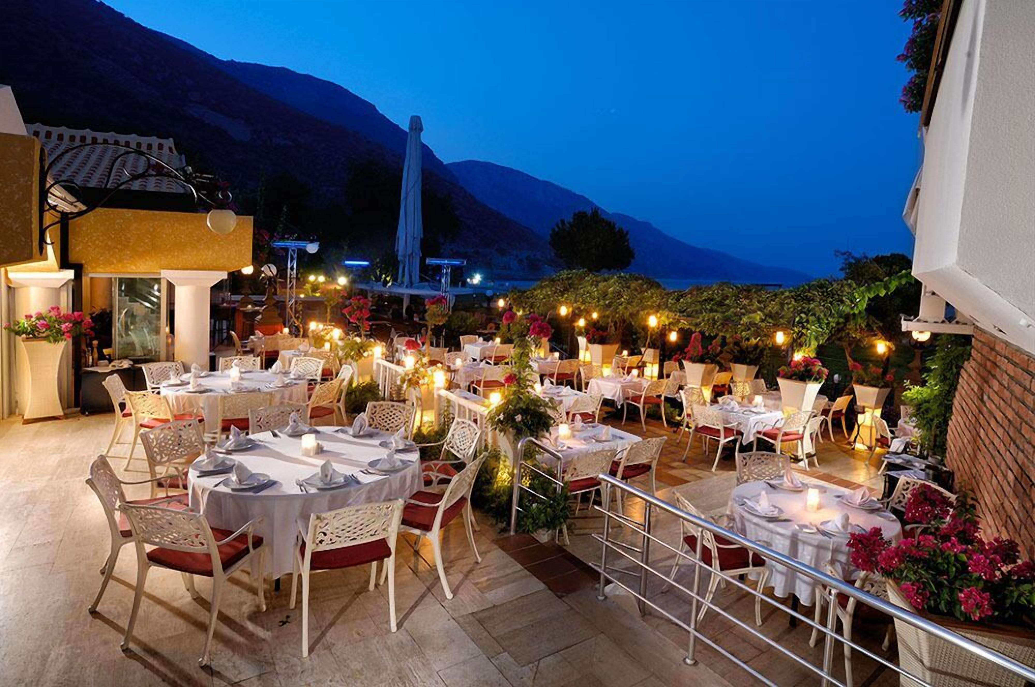 An outdoor dining area with round tables and chairs, surrounded by plants and decorative lights.