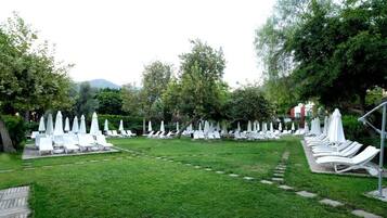 Un jardín con hileras de sillones blancos y sombrillas, rodeado de árboles y una montaña al fondo.