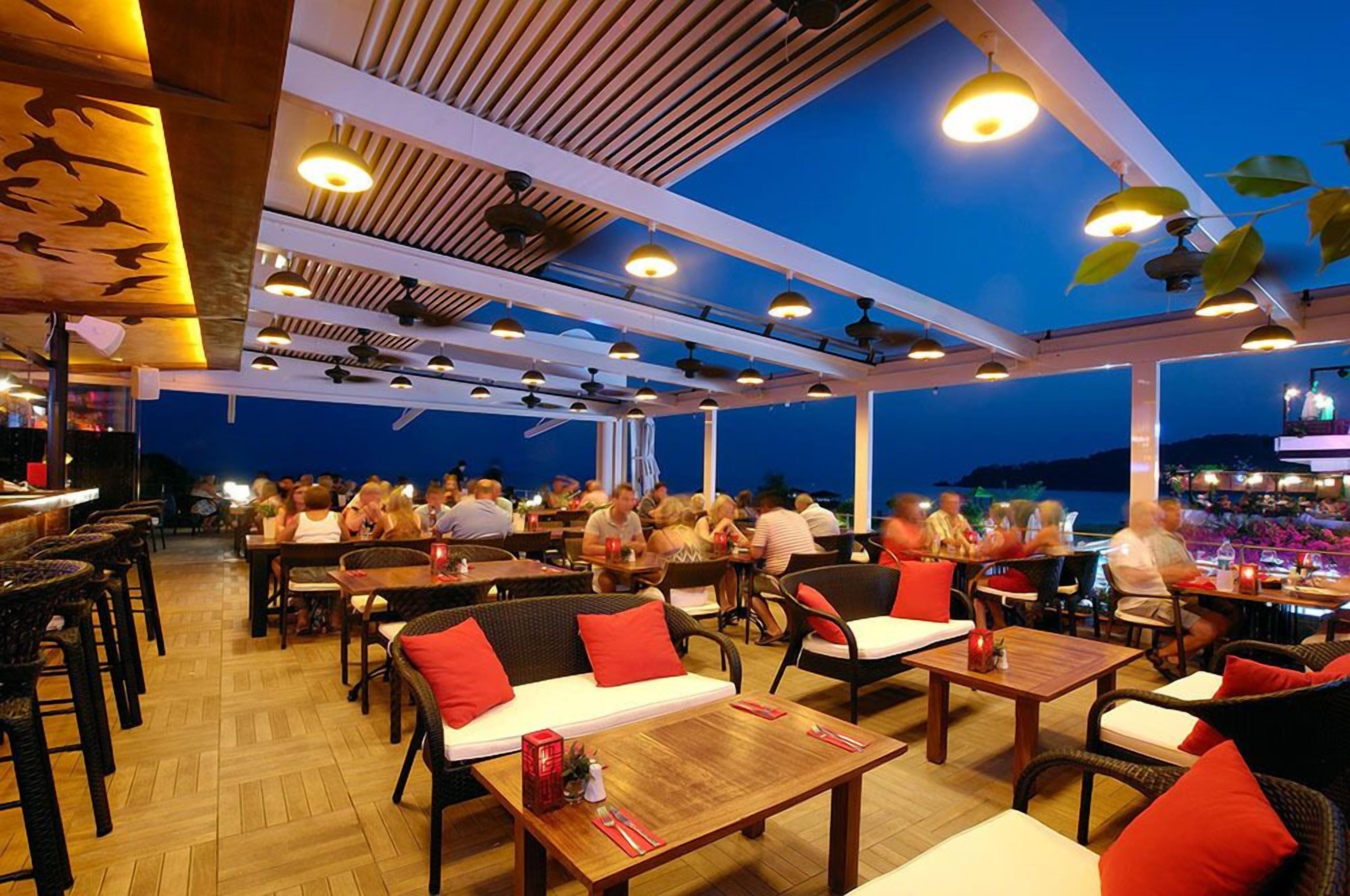 An outdoor restaurant with wooden tables and chairs, a covered seating area, and a view of the sea.