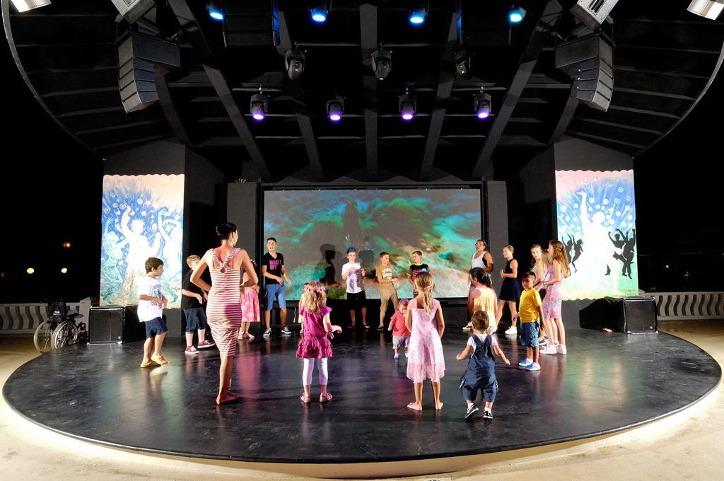 Um palco com um grupo de crianças e adultos, uma tela grande exibindo arte abstrata, e uma cadeira de rodas em primeiro plano.