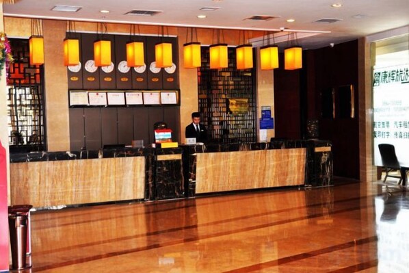 A hotel reception area with a wooden reception desk, a man standing behind it, and a wall with hanging lamps and framed items. - Huadong Hotel (Qitaihe (Xinjiang))