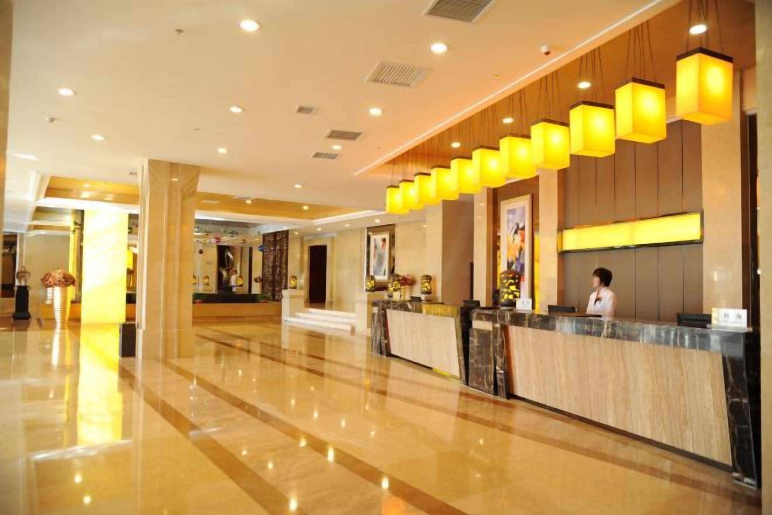A hotel lobby with a reception desk, a woman behind the desk, and a modern design with wooden panels and yellow lighting.