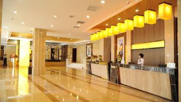 Un vestíbulo de hotel con una recepción, una recepcionista detrás del mostrador y un diseño moderno con paneles de madera e iluminación amarilla.
