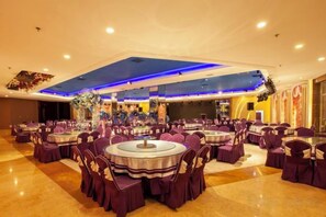 Un salón de banquetes con mesas y sillas redondas, un escenario con decoraciones florales e iluminación azul en el techo.