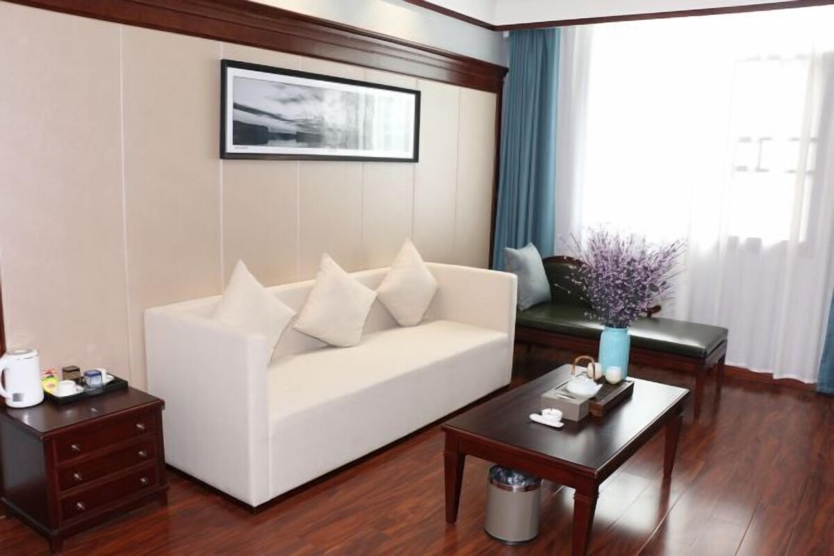 Ein modernes Hotelzimmer mit einem weißen Sofa, einem Holztisch, einem gerahmten Bild an der Wand und einem Fenster mit blauen Vorhängen.