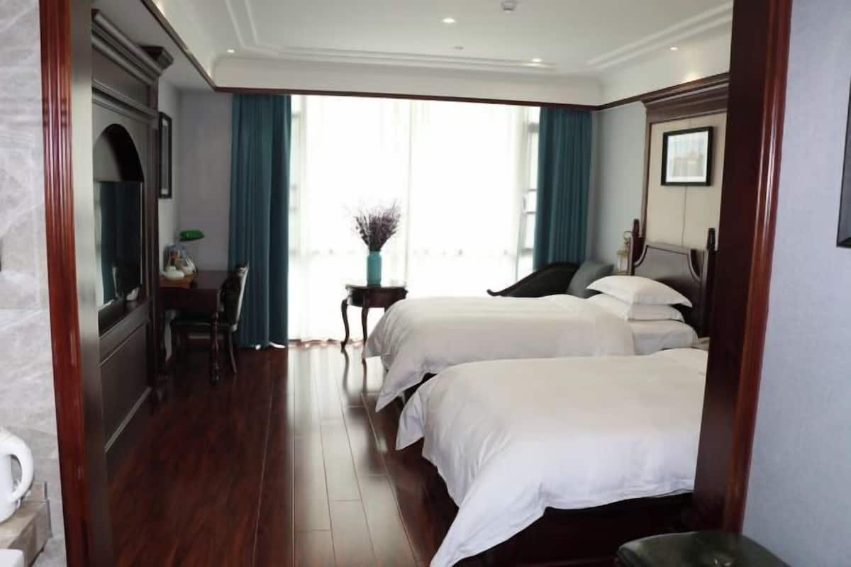 Habitación de hotel con dos camas, un escritorio, una silla, una mesita con un florero y un ventanal con cortinas.