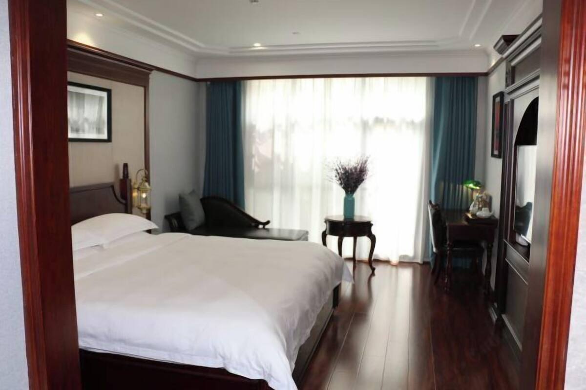 Habitación de hotel con una cama grande, un escritorio, una silla, una mesita con un jarrón y una televisión.