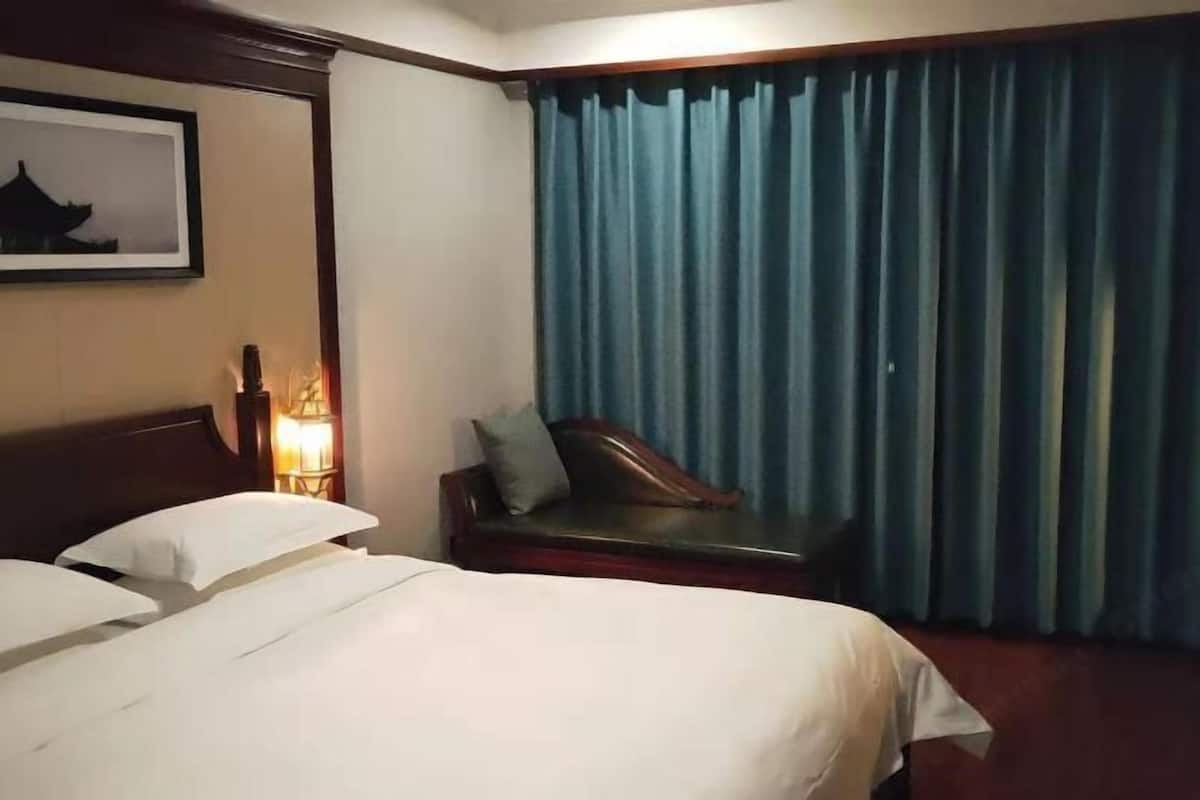 Habitación de hotel con cama, mesita de noche con lámpara, sofá y un ventanal con cortinas.