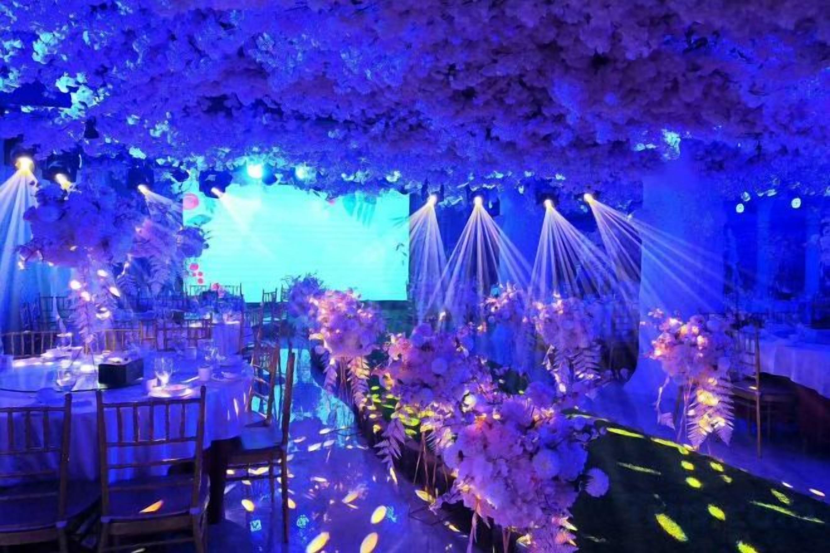An indoor venue with tables and chairs arranged for an event, decorated with floral arrangements and draped fabric.