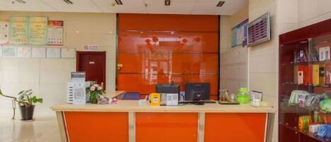 A reception desk with a computer, a telephone, and a counter with various items. There are signs on the wall and a plant on the left.