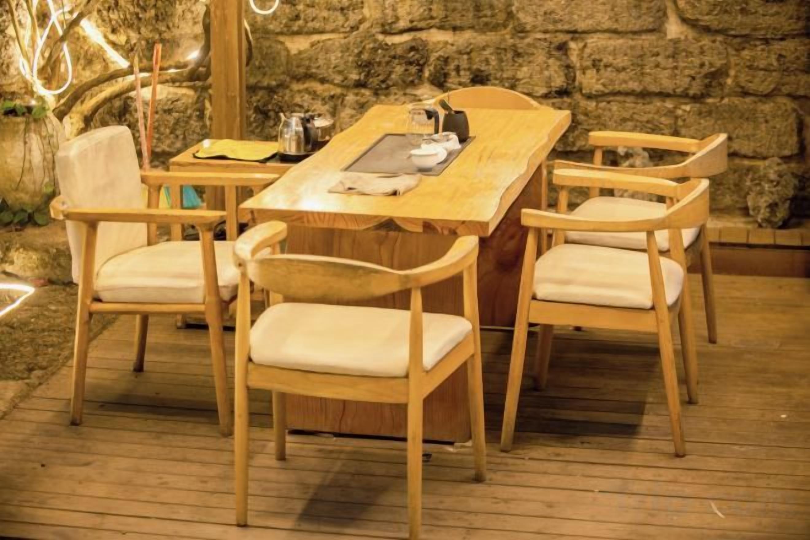 A wooden dining table with four chairs, set on a wooden deck with a stone wall in the background.