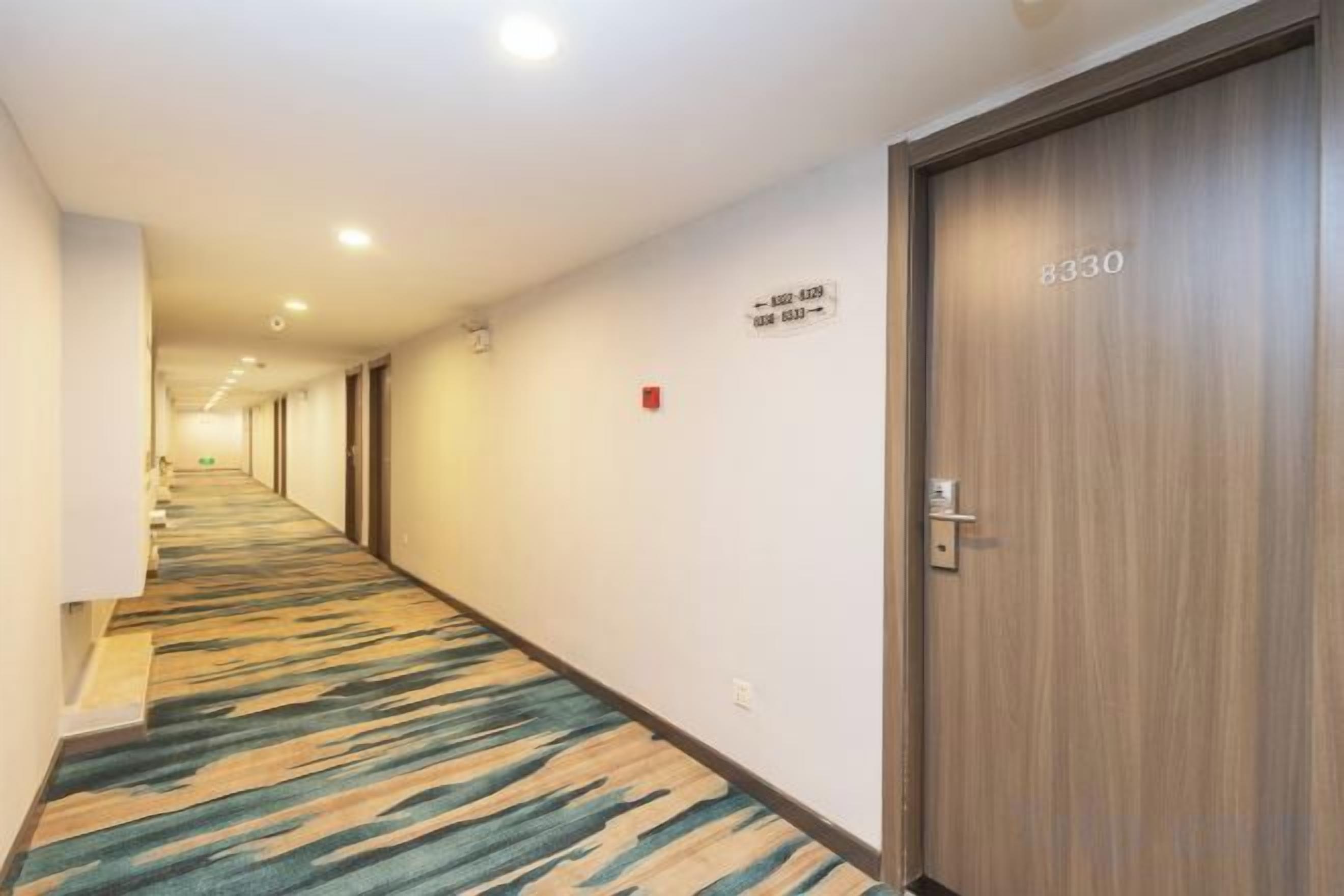 A hotel corridor with carpeted flooring, a wooden door with a number, and a fire alarm on the wall.