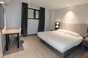 A hotel room with a bed, a desk, a chair, a nightstand, and a lamp.